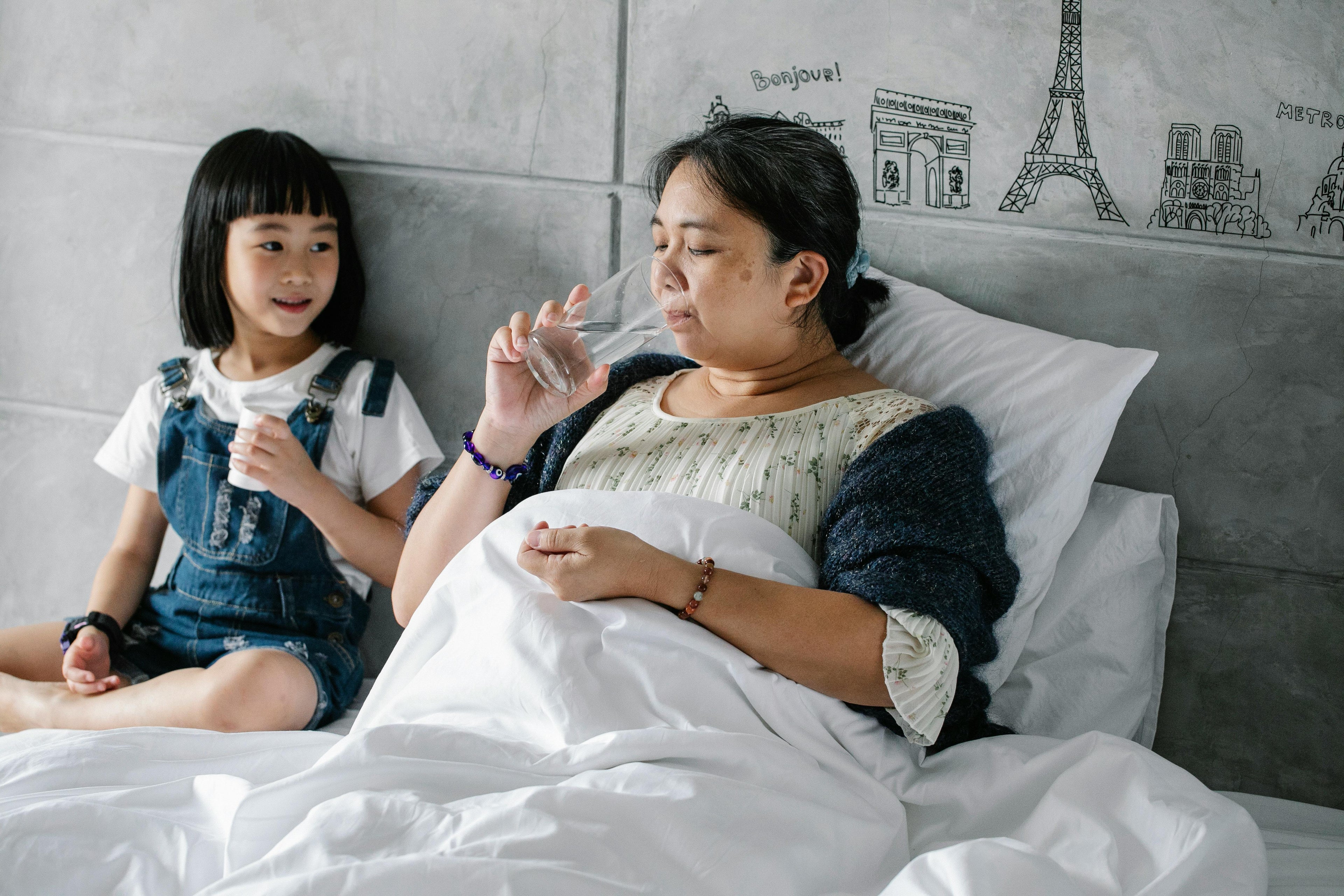 Woman in bed with a child holding a glass of water, with a gray wall and decorative elements in the background.