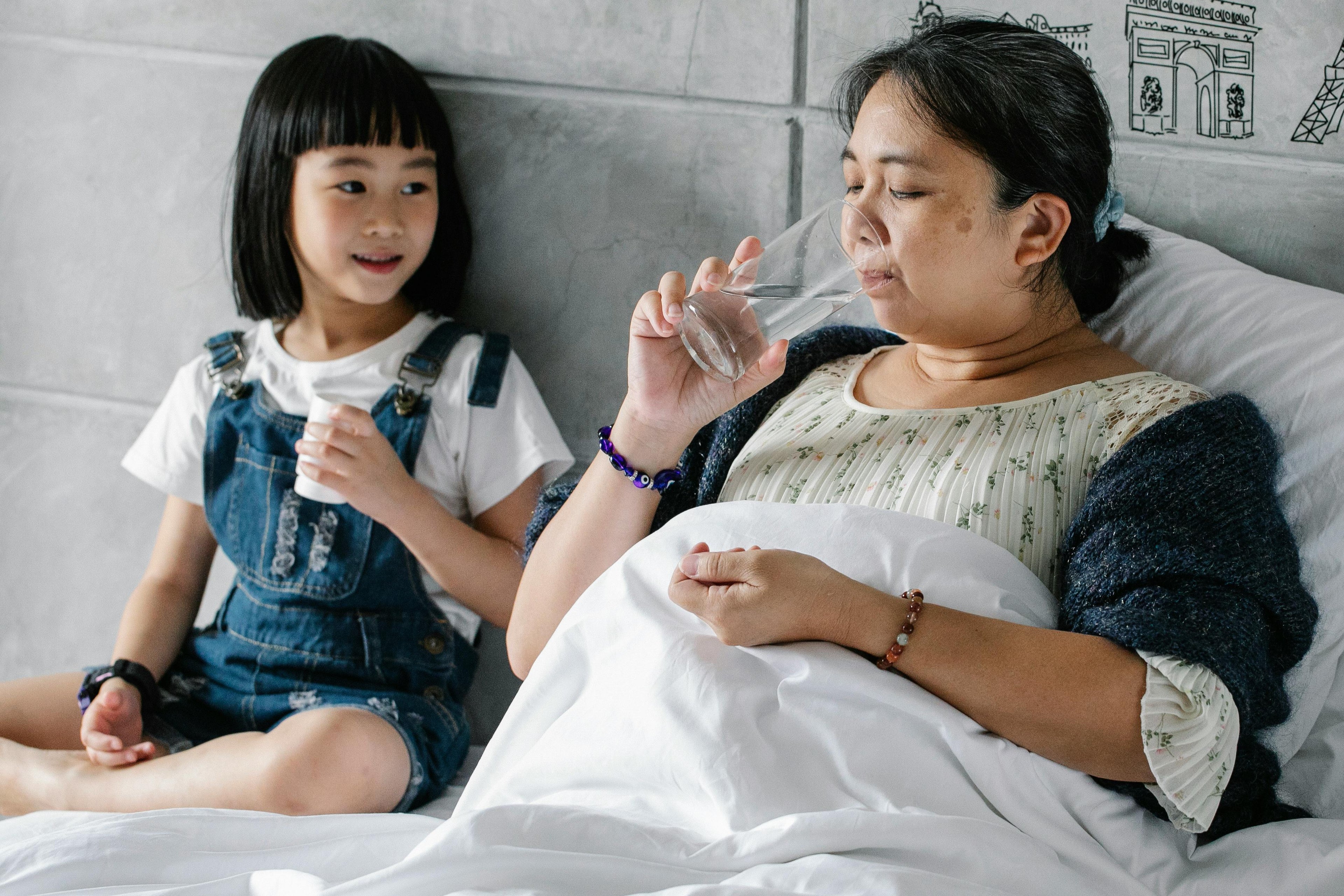 Woman in bed with a child holding a glass of water, with a gray wall and decorative elements in the background.